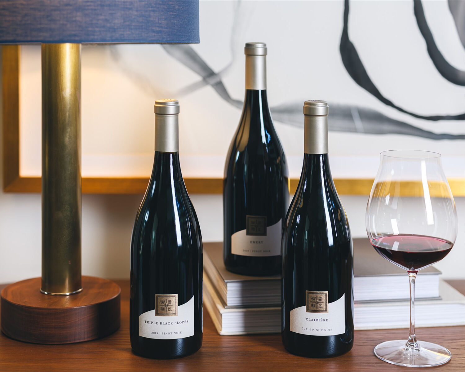 Three bottles of wine laying on wooden furniture next to books, a lamp and a glass of red wine.